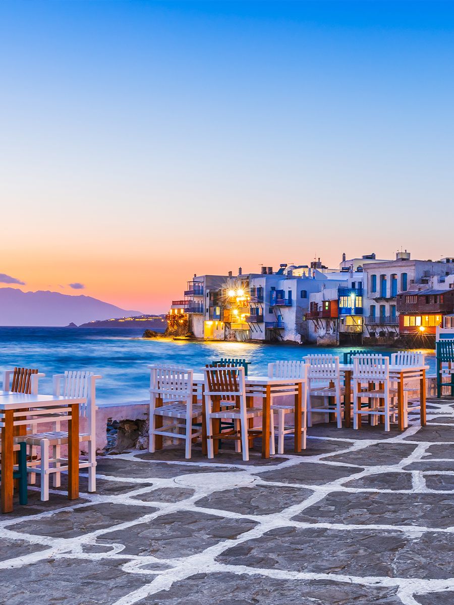 Seaside view with white and wooden chairs by a stone path, vibrant sunset sky, and white buildings along the coastline.