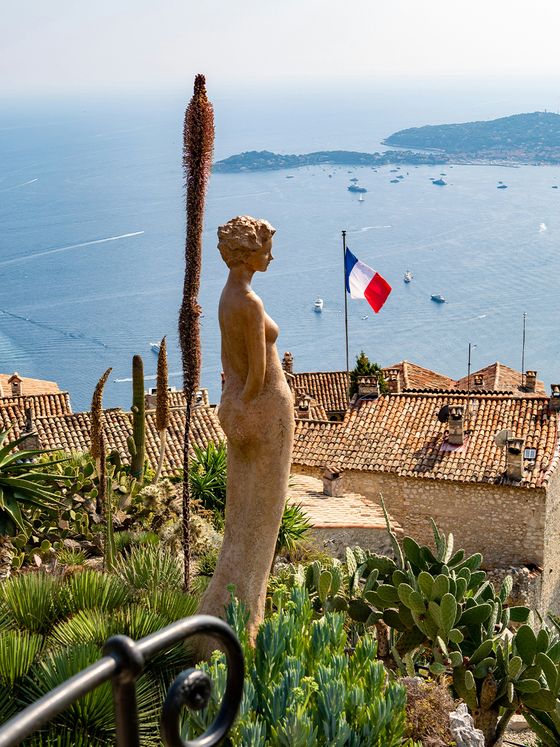A stone statue overlooks a coastal village with terra-cotta rooftops, cacti, a French flag, and a view of the sea with boats.