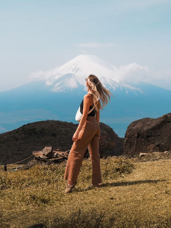 Woman with long hair stands on grassy hill, gazing at a distant snow-capped mountain under a clear blue sky.