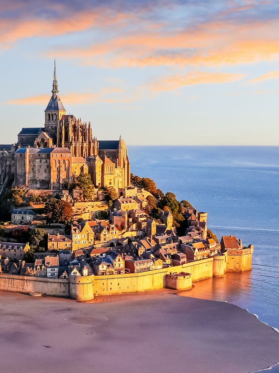 Mont-Saint-Michel at sunset, with its towering abbey and historic buildings, surrounded by sandy tidal flats and a calm blue sea.