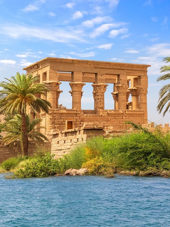 Ancient Egyptian stone temple with columns near a river, surrounded by palm trees and greenery under a partly cloudy blue sky.