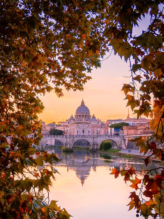 St. Peter's Basilica and bridge at sunset, framed by autumn leaves reflecting on a calm river.
