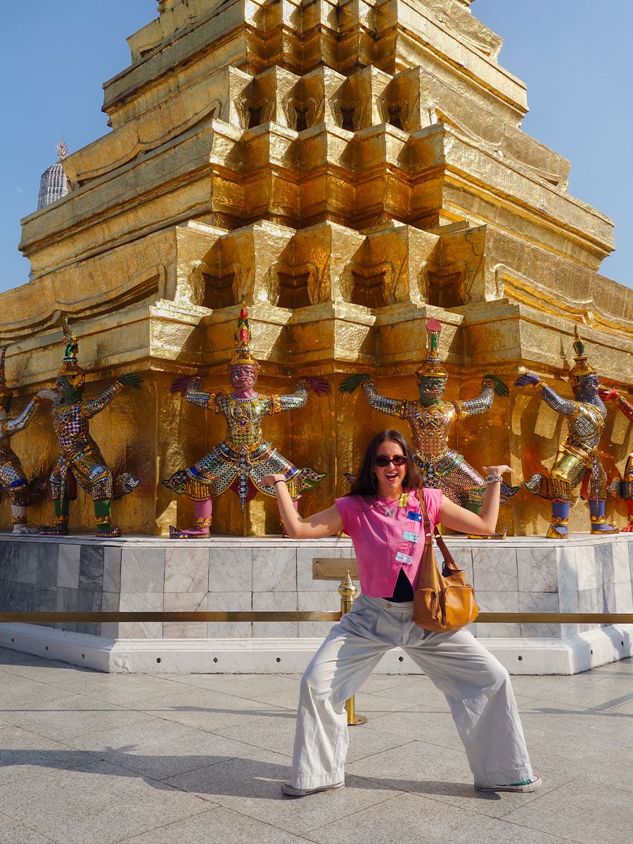 Woman in a pink shirt posing playfully in front of a golden temple with colorful statues, under a clear blue sky.