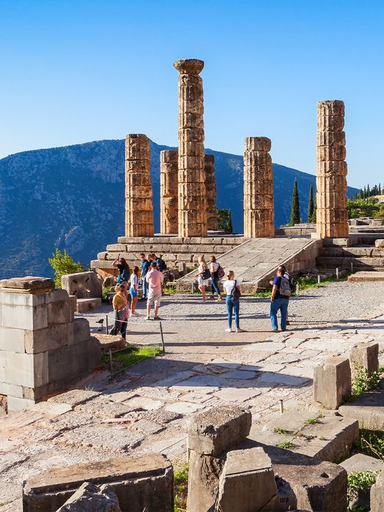 Visitors explore ancient ruins with tall stone columns against a mountain backdrop and clear blue sky.
