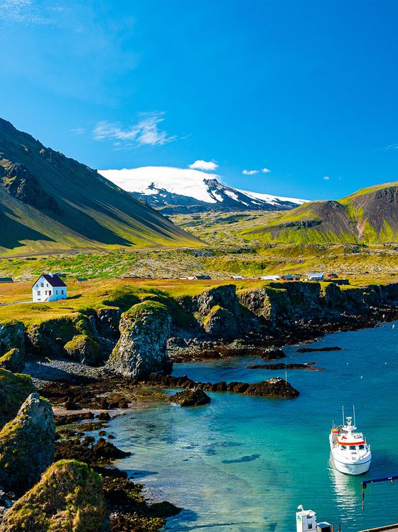 A scenic coastal landscape with a small white house, rocky cliffs, clear blue water, a docked boat, and a snow-capped mountain in the background.