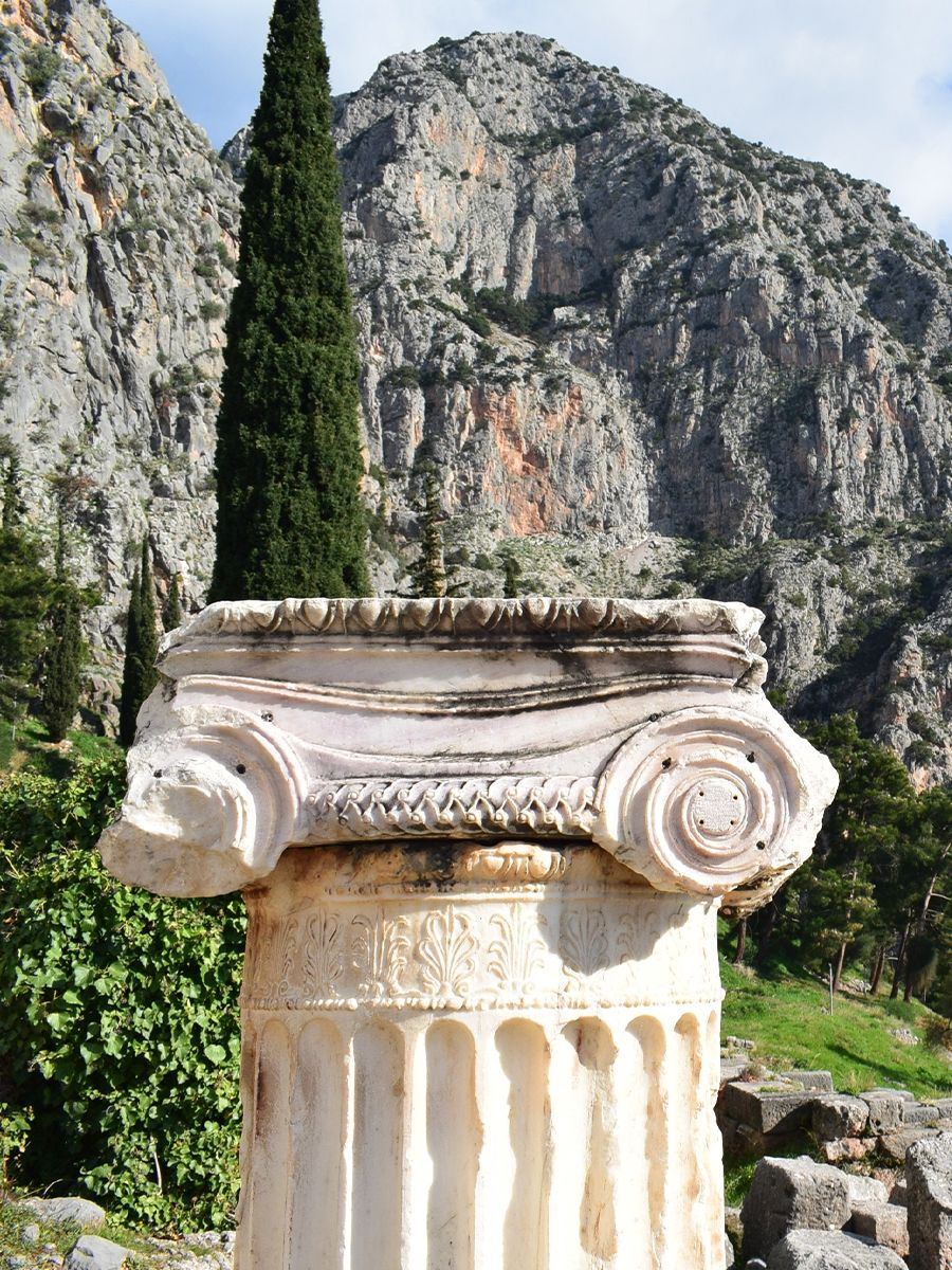 A weathered Ionic column capital with intricate carvings set against a backdrop of rugged mountains and lush greenery.