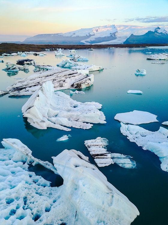 Icebergs floating in a serene, turquoise glacial lagoon with snow-capped mountains in the distance under a pastel sky.