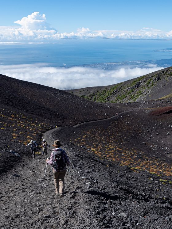 Hikers with trekking poles descend a rocky trail on a volcanic landscape with a distant view of the sea and scattered clouds below.