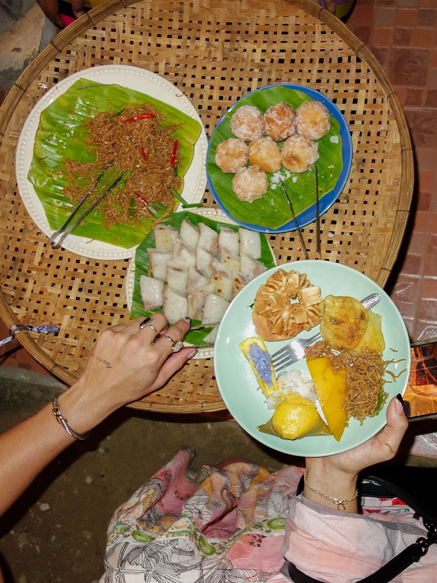 A wooden table with assorted Southeast Asian foods, including noodles, rolls, and desserts on plates. Two hands are reaching for the dishes.