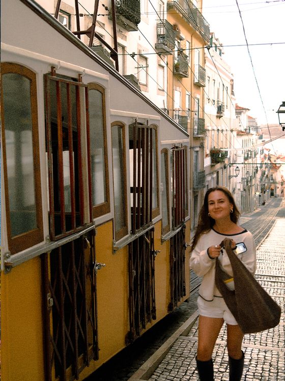 A woman walks uphill beside a vintage yellow tram on a cobblestone street in a European city, holding a brown bag and smiling.