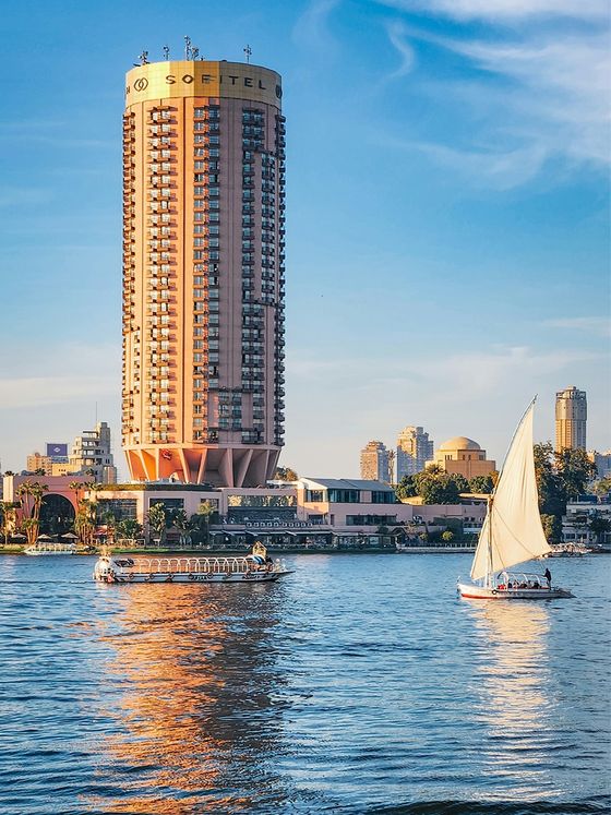 A tall cylindrical hotel building by a river with a sailboat and a motorboat under a clear blue sky.