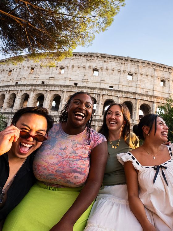 Four people laugh and sit together in front of the Colosseum, with trees framing the scene, under a clear blue sky.