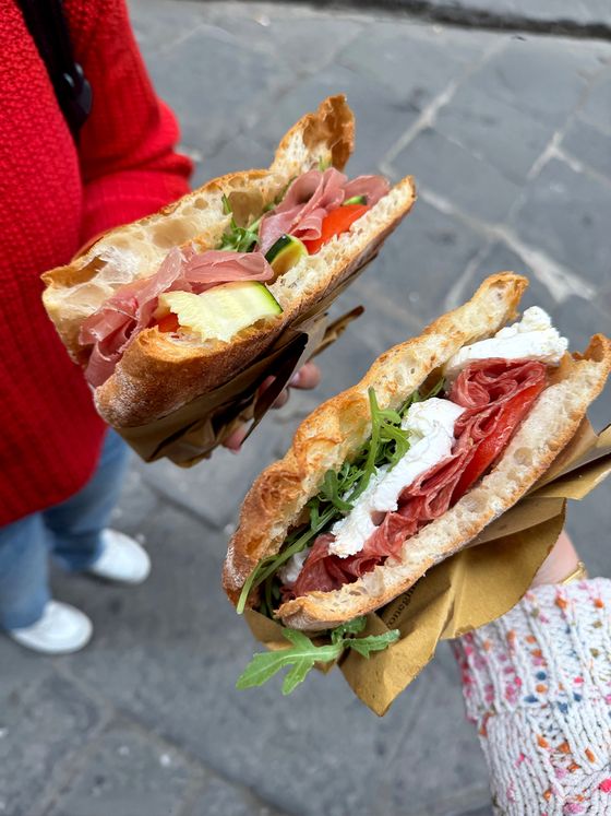 Two hands hold sandwiches filled with prosciutto, arugula, and cheese on crusty bread, wrapped in paper, against a street background.