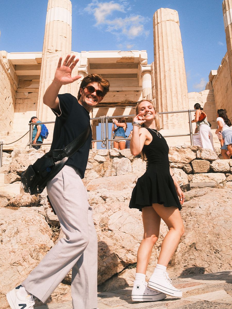 Two people smiling and waving, walking in front of ancient ruins with columns, surrounded by a clear blue sky and tourists.