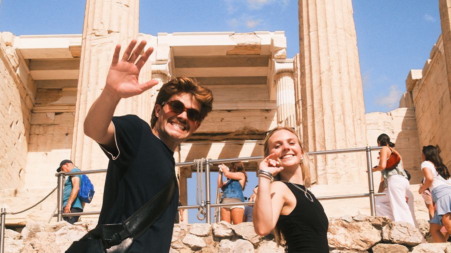 Two people smiling and waving, walking in front of ancient ruins with columns, surrounded by a clear blue sky and tourists.