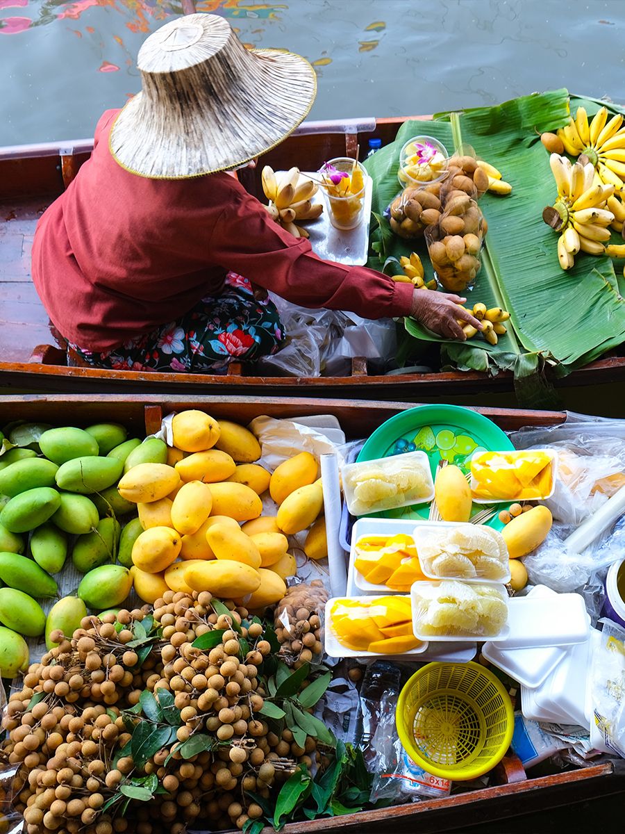 A vendor in a straw hat sells various tropical fruits, including mangoes and longans, from a boat at a floating market.