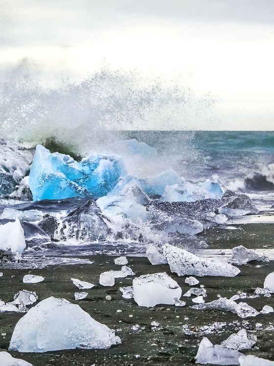 Waves crashing onto large blue ice chunks scattered along a dark sandy beach under a cloudy sky.