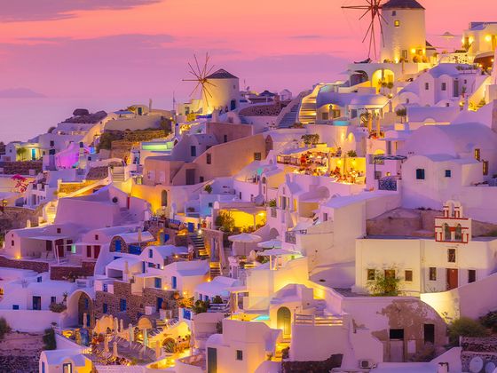 A picturesque view of Santorini during sunset, with white buildings, illuminated windmills, and a pink-orange sky in the background.