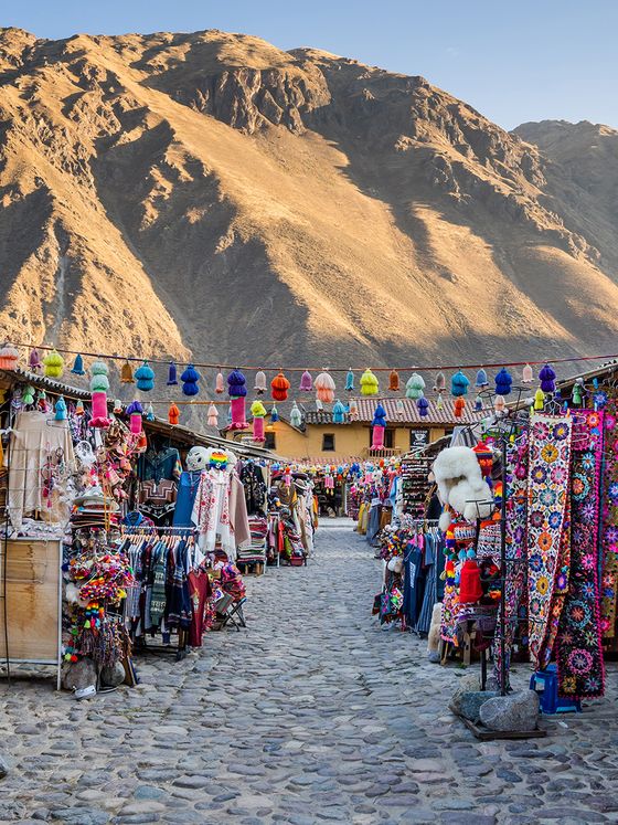 A vibrant market with colorful textiles and crafts lining a cobblestone path, set against a backdrop of majestic mountains in the early evening light.