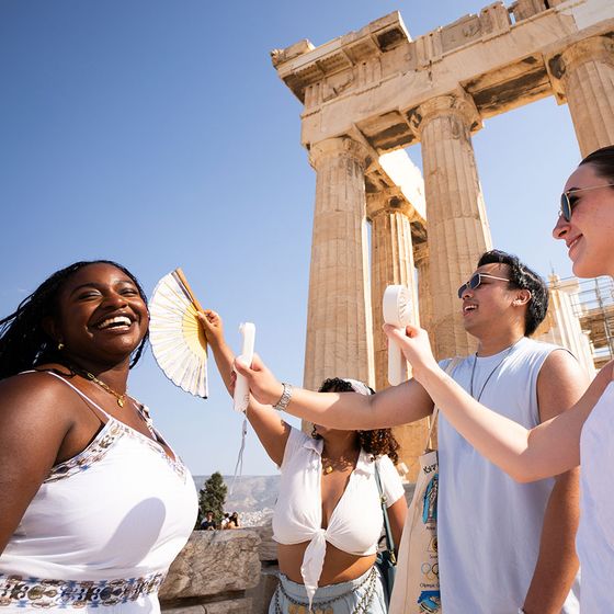Four people enjoying a sunny day, fanning each other in front of ancient Greek columns, smiling and wearing summer attire.