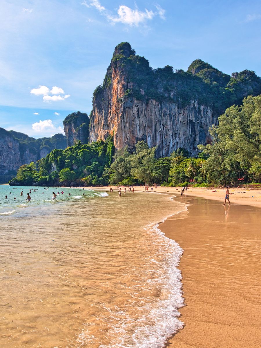 Sandy beach with people enjoying the water, surrounded by lush greenery and towering limestone cliffs under a clear blue sky.