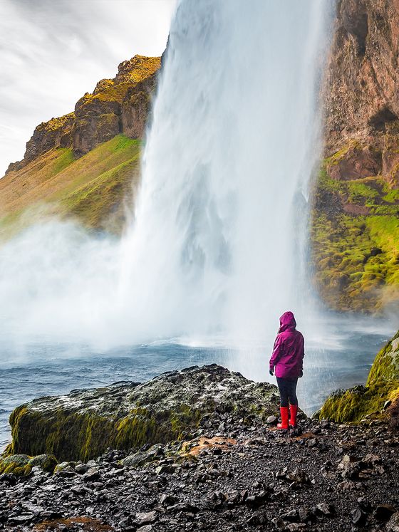 Person in purple jacket and red boots stands on rocky terrain, gazing at a tall, misting waterfall surrounded by mossy cliffs.