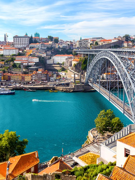 Scenic view of a historic iron bridge spanning a blue river, with colorful buildings and lush hills under a clear sky.