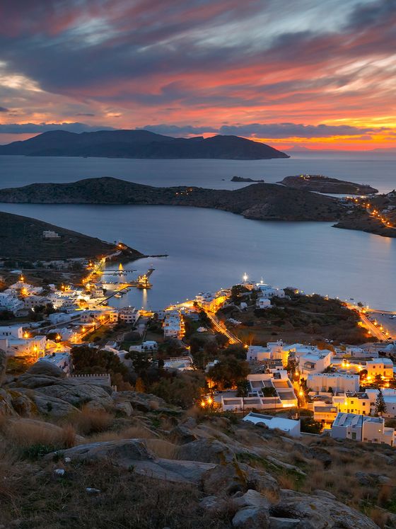 A coastal town glows with lights at dusk, under a vibrant sunset sky with island silhouettes in the background.