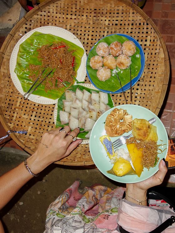 A person's hand holding a plate of assorted traditional snacks, including banana, sticky rice, and pastries, over a woven table with more dishes.