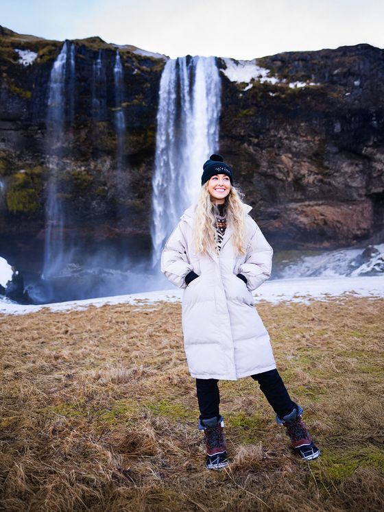 A person in a white coat and beanie stands smiling in front of a tall waterfall with mist, on a grassy area.