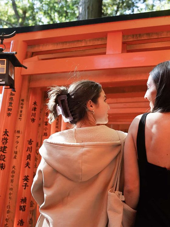 Two women walk through a path lined with vibrant orange torii gates, with sunlight filtering through above.