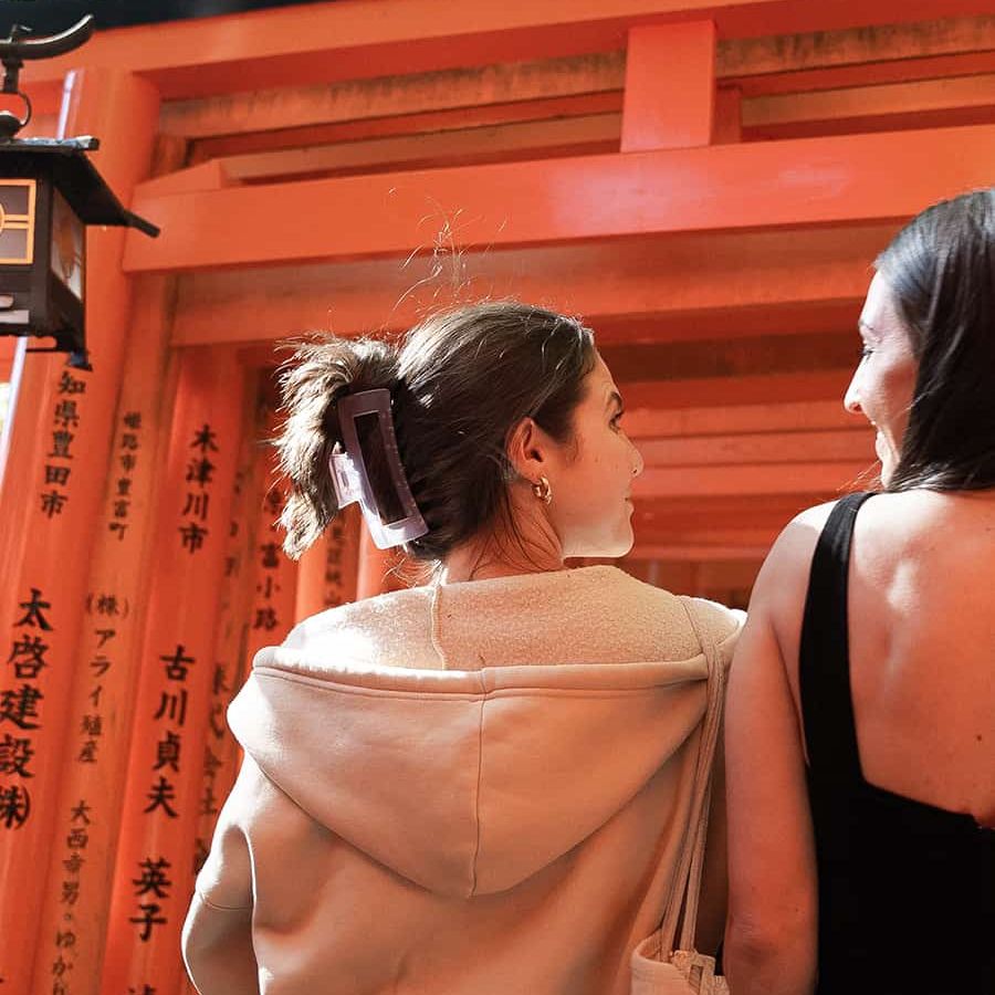 Two women walk through a path lined with vibrant orange torii gates, with sunlight filtering through above.