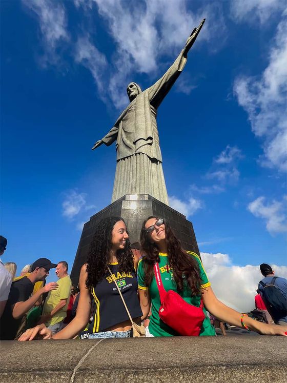 Two women smiling in front of the Christ the Redeemer statue against a blue sky with clouds.