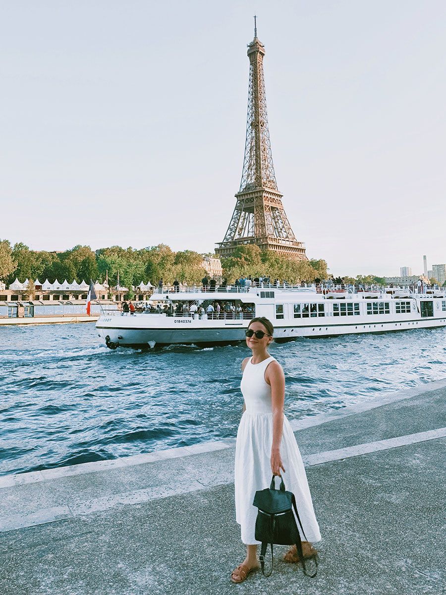 A woman in a white dress stands by the Seine River, with the Eiffel Tower in the background and a boat passing by.