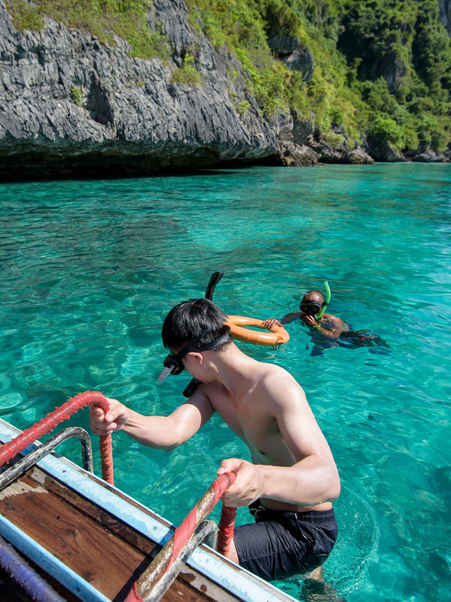 A person climbs a boat ladder from clear turquoise water, while another person snorkels nearby against a backdrop of rocky cliffs and greenery.