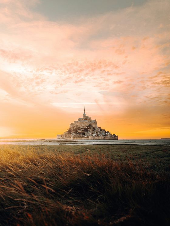 Mont Saint-Michel at sunset, with a vibrant orange sky and foreground grass gently swaying in the breeze.