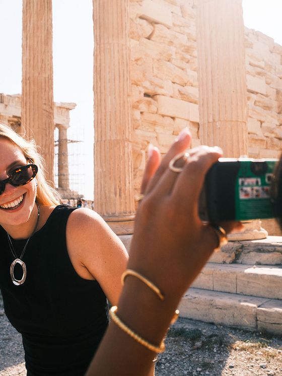 Woman in sunglasses smiling, taking a photo at an ancient stone temple with columns, under bright sunlight.