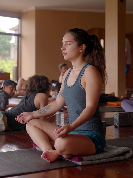 A woman sits cross-legged on a yoga mat, meditating with eyes closed in a room with other people practicing yoga.