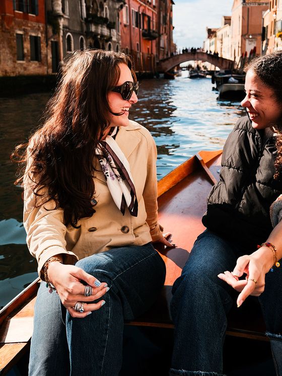 Two friends laughing in a small wooden boat on a sunlit Venetian canal, colorful buildings and arched bridge in the background.