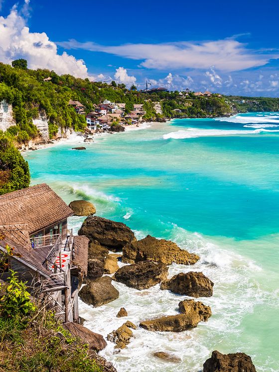 Scenic view of a coastline with turquoise waters, rocky cliffs, lush greenery, and beachside huts under a bright blue sky.
