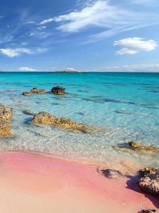 Scenic view of a pink sandy beach with rocky outcrops, clear turquoise water, and a bright blue sky with wispy clouds.