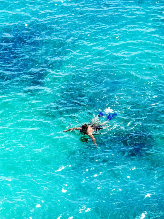 Person swimming in clear turquoise ocean water, creating small ripples. Shadows from underwater features are visible.
