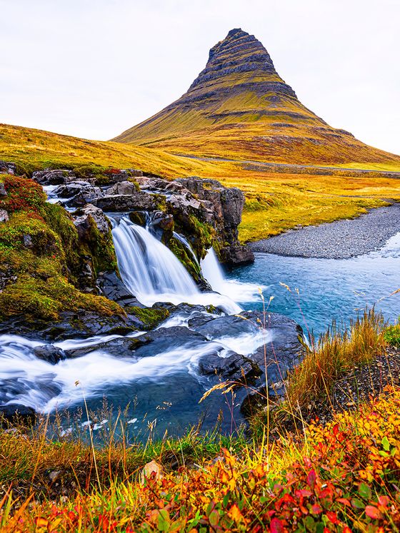 A scenic view of Kirkjufell mountain with a flowing waterfall, surrounded by autumn foliage and a clear stream in Iceland.