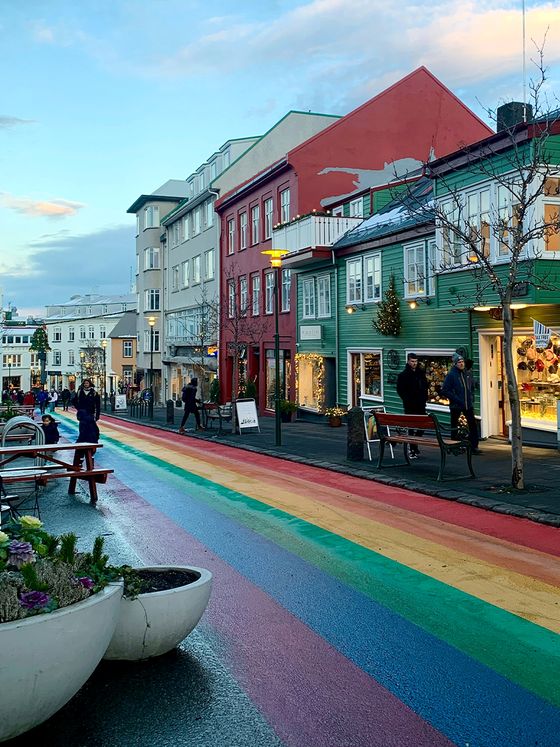 A colorful street with a rainbow-painted pathway, lined with vibrant buildings, shops, and pedestrians under a cloudy blue sky.