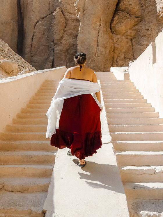 A woman in a red dress and white shawl walks up a stone staircase between rocky cliffs on a sunny day.