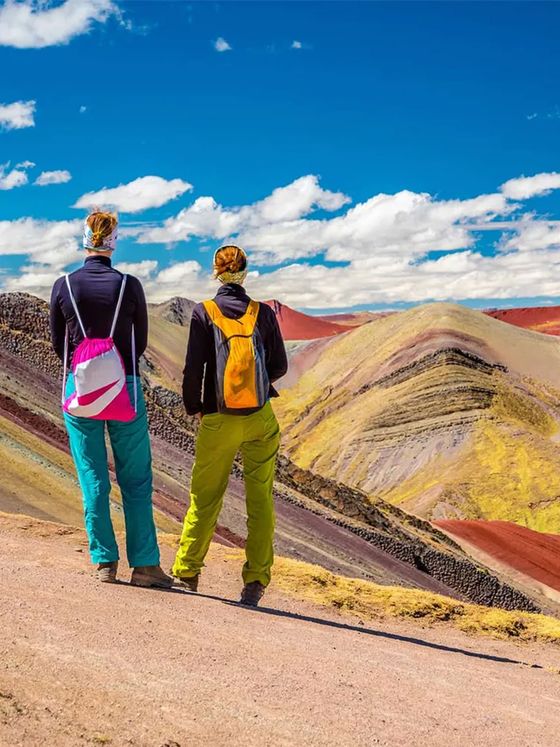 Two hikers with backpacks admire colorful, layered mountains under a blue sky with clouds. The vibrant terrain features shades of red, yellow, and purple.