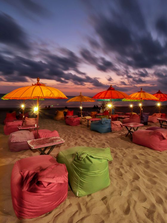 Vibrant bean bags and colorful umbrellas on a sandy beach at sunset, with a dramatic cloudy sky in the background.