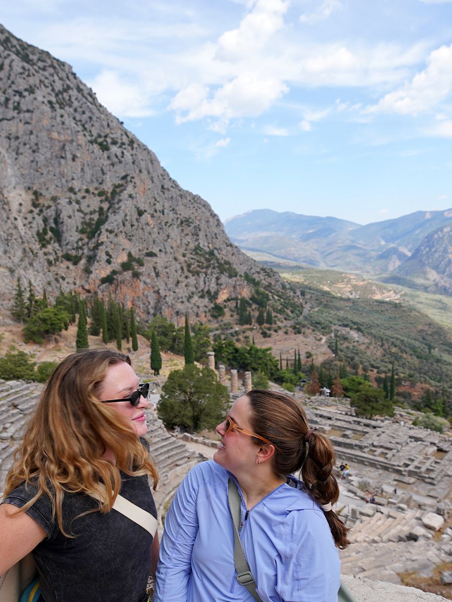 Two people smiling at each other with ancient ruins, trees, and mountains in the background under a partly cloudy sky.