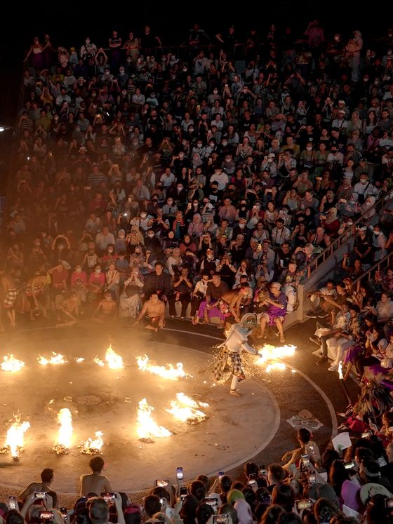 A large crowd gathers in a circular arena to watch a fire performance at night, with flames surrounding the performer in the center.