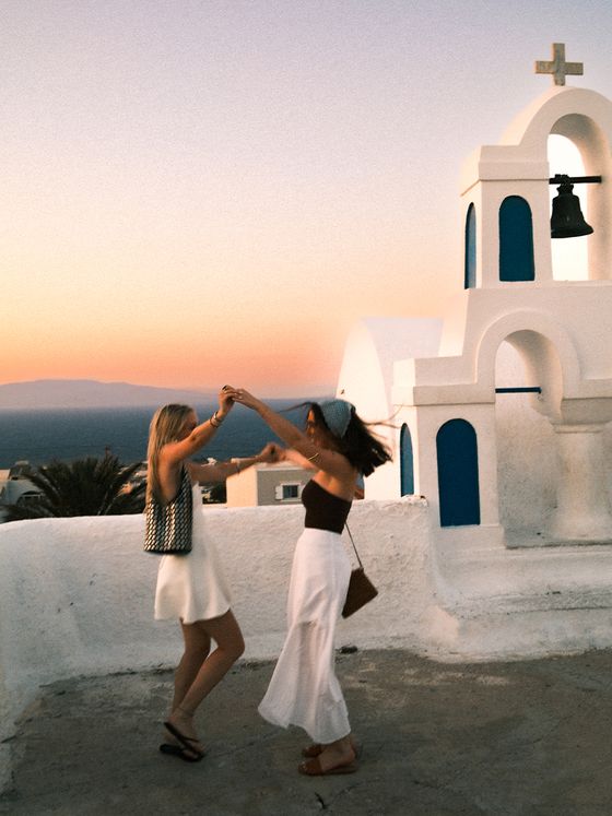 Two people dance joyfully on a rooftop at sunset, with a white church and bell tower in the background, against a colorful sky.
